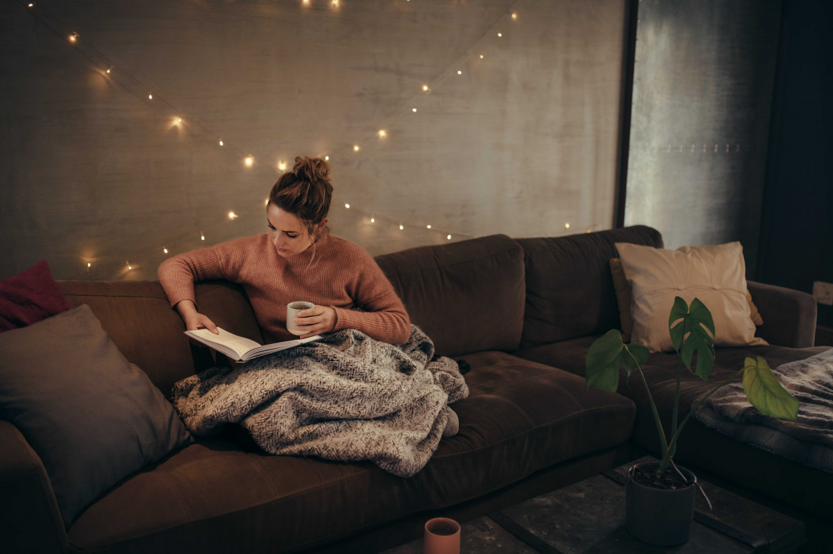 Paní si čte knížku na gauči s dekou a čajem v hygge atmosféře